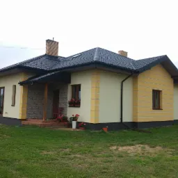 Widok z zewnątrz na dom jednorodzinny z nową, ciemną dachówką, kremowo-żółtą elewacją z dekoracyjnymi pasami i ceglanym kominem, otoczony zielonym trawnikiem i kwiatami w donicach.