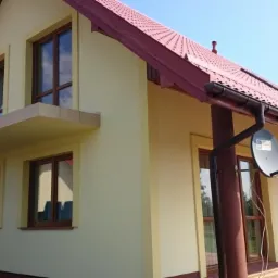 Żółty dom jednorodzinny z brązowymi oknami i czerwoną dachówką, widoczny z zewnątrz w słoneczny dzień, z zamontowaną anteną satelitarną.