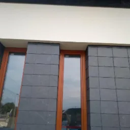 Fragment elewacji budynku z oknami, wykończony ciemnoszarymi płytkami imitującymi kamień, z widocznym otworem wentylacyjnym w ścianie.