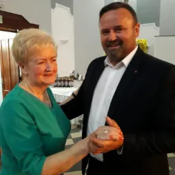 Uśmiechnięty mężczyzna w czarnym garniturze i białą koszulą tańczy z kobietą w zielonej sukience na tle sali z zastawionym stołem i biało-szarą ścianą.