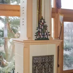 Biały piec kaflowy z ażurowymi drzwiczkami, ozdobiony małą choinką z figurkami, na tle okna z widokiem na ogród.