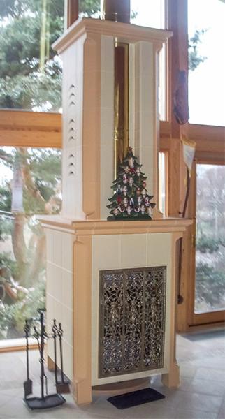 Biały piec kaflowy z ażurowymi drzwiczkami, ozdobiony małą choinką z figurkami, na tle okna z widokiem na ogród.