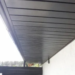 Ciemnoszare panele podbitki dachowej z widocznym systemem rynnowym, zamontowane na białej elewacji budynku.