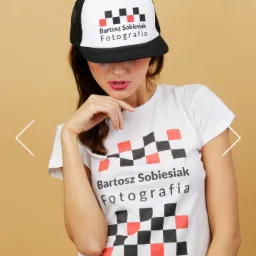 Kobieta w czapce trucker i koszulce z logo fotografa, pozująca na jednolitym, beżowym tle.