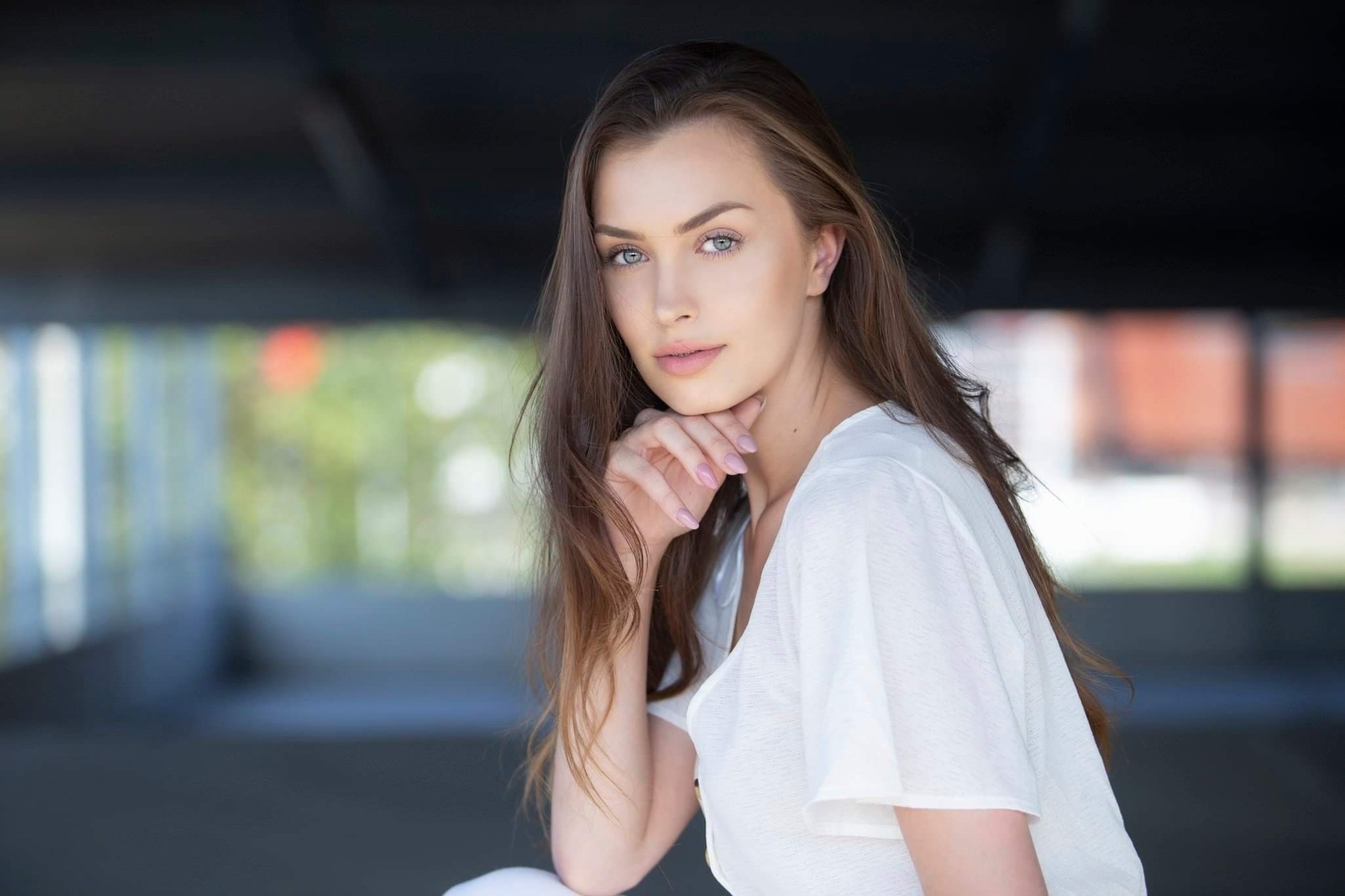 Portret młodej kobiety z długimi brązowymi włosami, opierającej podbródek na dłoni, w białej bluzce na tle rozmytej przestrzeni miejskiej.