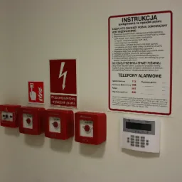 Rząd czerwonych, kwadratowych przycisków alarmowych z napisem 'Wyłącznik Główny' na ścianie, obok instrukcja postępowania na wypadek pożaru i panel sterowania alarmem.