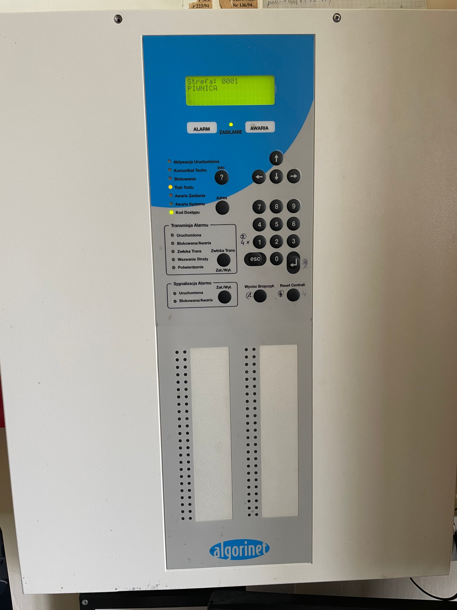 Panel sterowania systemem alarmowym Algorinet z wyświetlaczem wskazującym 'Strefa: 0001 PIWNICA', przyciski numeryczne i funkcyjne, diody sygnalizacyjne Alarm i Awaria. Widoczne otwory na dodatkowe...