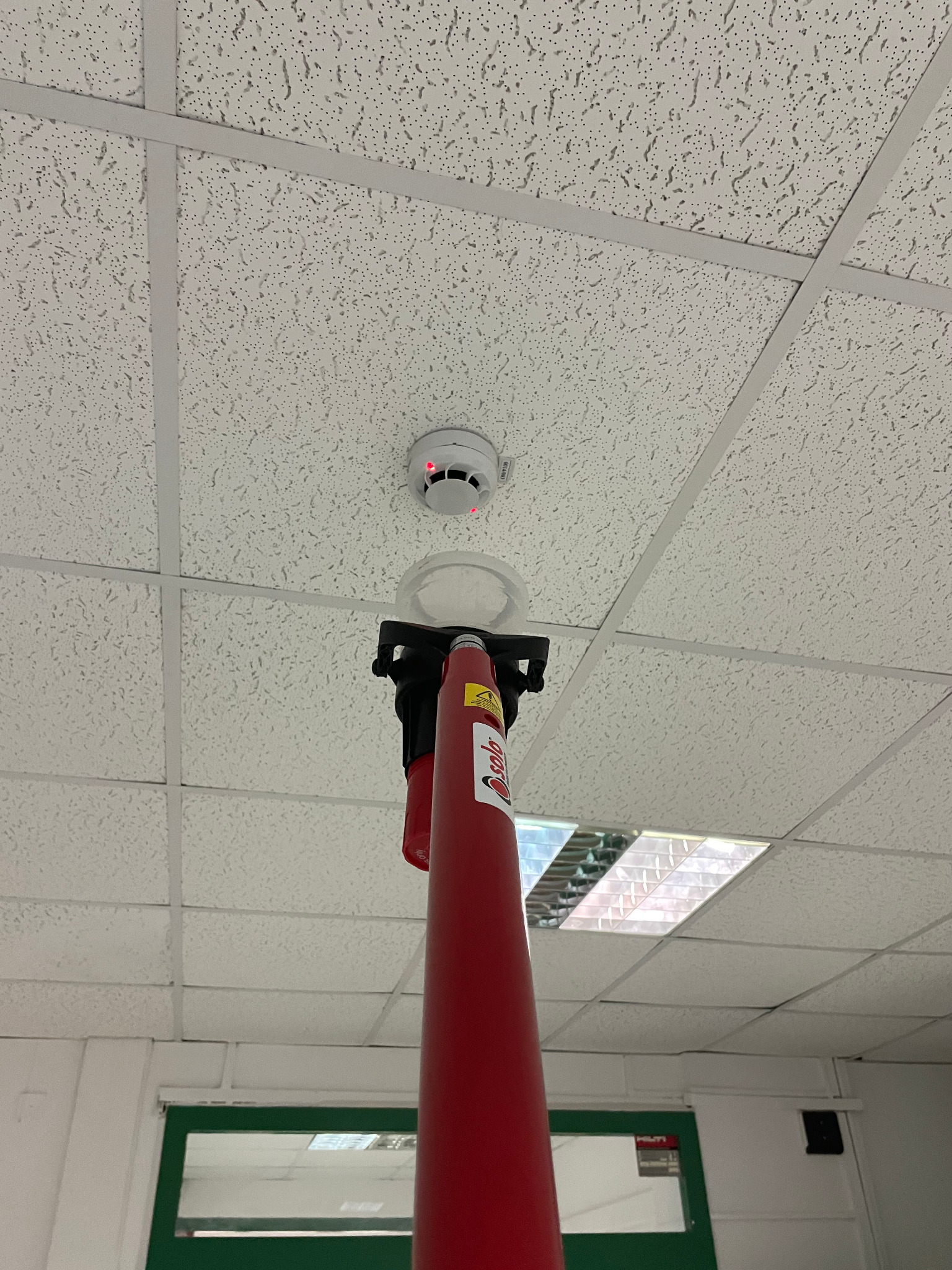 Testowanie czujnika dymu na suficie podwieszanym za pomocą specjalistycznego testera na wysięgniku; widoczne czerwone diody na czujniku.