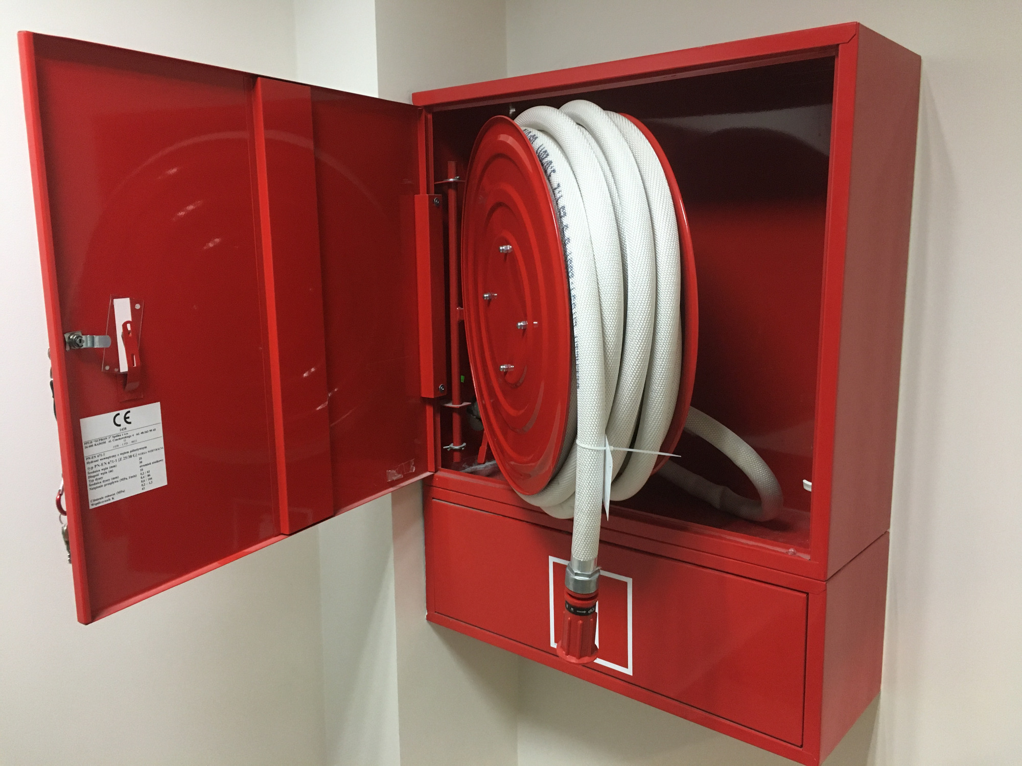Czerwona szafka hydrantowa z otwartymi drzwiami, zawierająca zwinięty wąż strażacki z dyszą, zamontowana na jasnej ścianie.