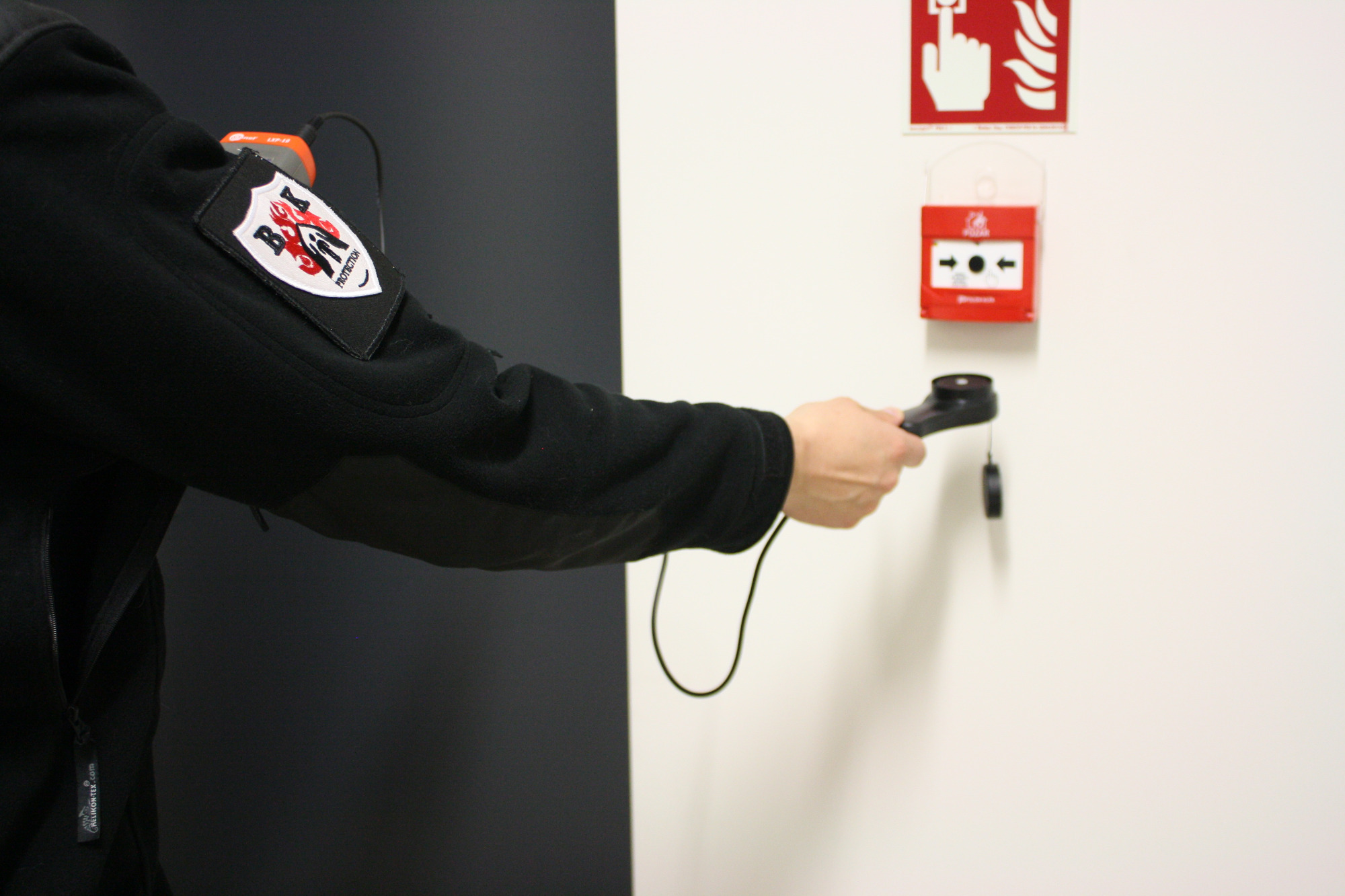 Osoba w czarnej bluzie z naszywką firmy dokonuje pomiaru czujnikiem dymu przyciskowego alarmu pożarowego na białej ścianie, obok znaku ewakuacyjnego.