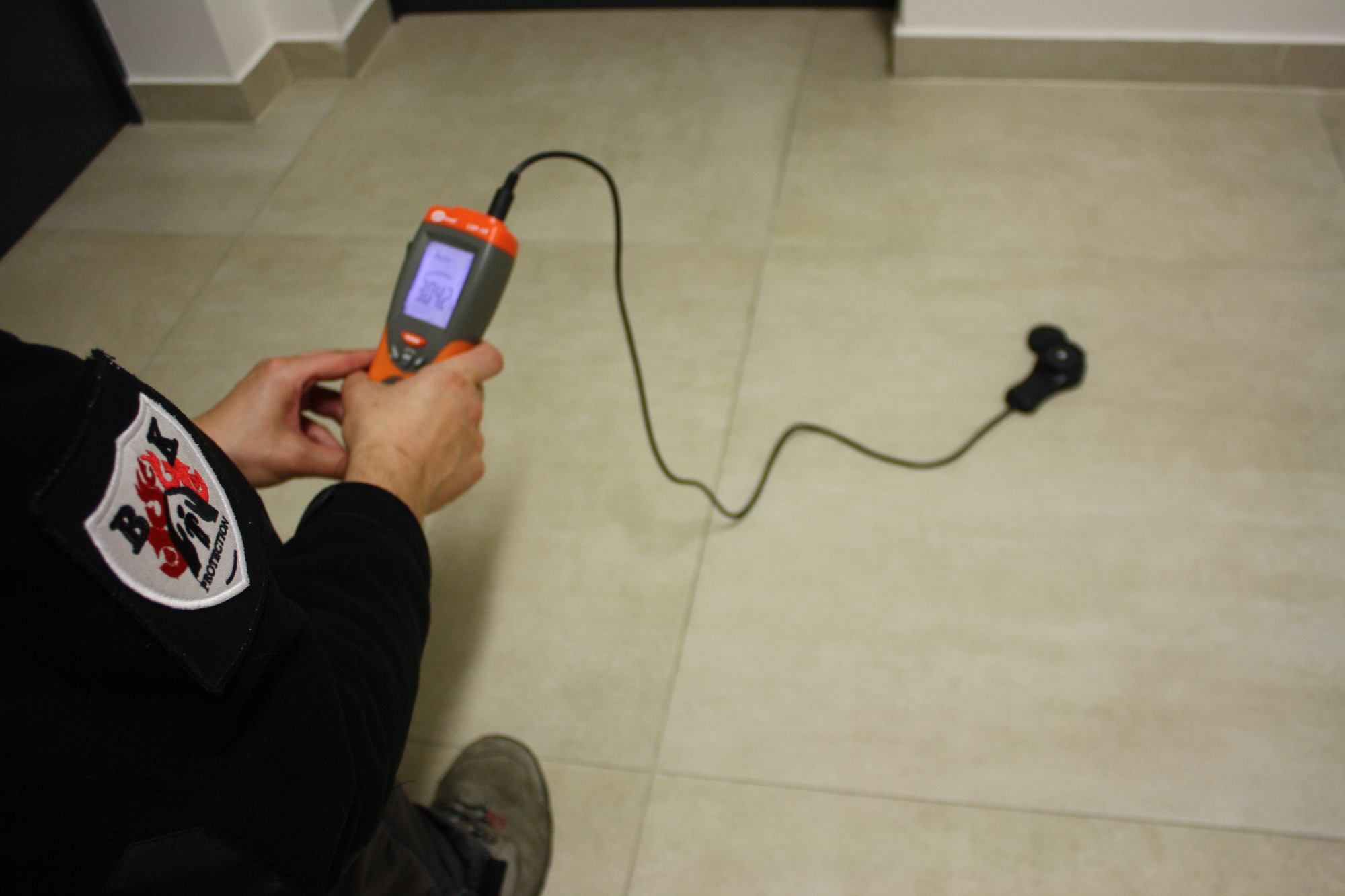 Osoba w uniformie z naszywką B&K Protection mierzy poziom dźwięku detektorem z wyświetlaczem LCD na kafelkowej podłodze, w pobliżu ściany.