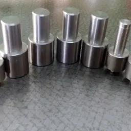 Siedem metalowych, cylindrycznych elementów o różnych średnicach, ułożonych na metalowej powierzchni z widocznymi śladami obróbki.