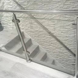 Nowoczesne schody z szarą okładziną i szkłem hartowanym, zabezpieczone stalową konstrukcją i poręczą, na tle ściany z dekoracyjną imitacją kamienia.