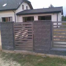 Nowoczesna brama wjazdowa z metalowymi przęsłami i kamiennymi słupkami na tle domu z ciemnym dachem.