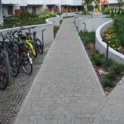 Szary chodnik z kostki brukowej prowadzący wzdłuż nowoczesnego budynku mieszkalnego z balkonami, widoczne rowery i elementy zieleni miejskiej.