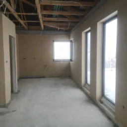 Wnętrze surowego domu po nałożeniu beżowych tynków, widoczne okna z widokiem na zimowy krajobraz, surowy betonowy podkład i odsłonięta drewniana konstrukcja dachu z widocznymi przewodami.