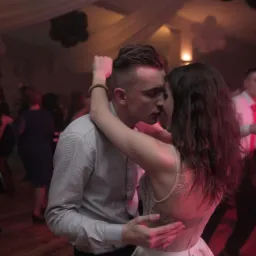 Para młoda w objęciach podczas pierwszego tańca na sali balowej z parkietem i innymi gośćmi w tle, oświetlenie sceniczne w odcieniach czerwieni.