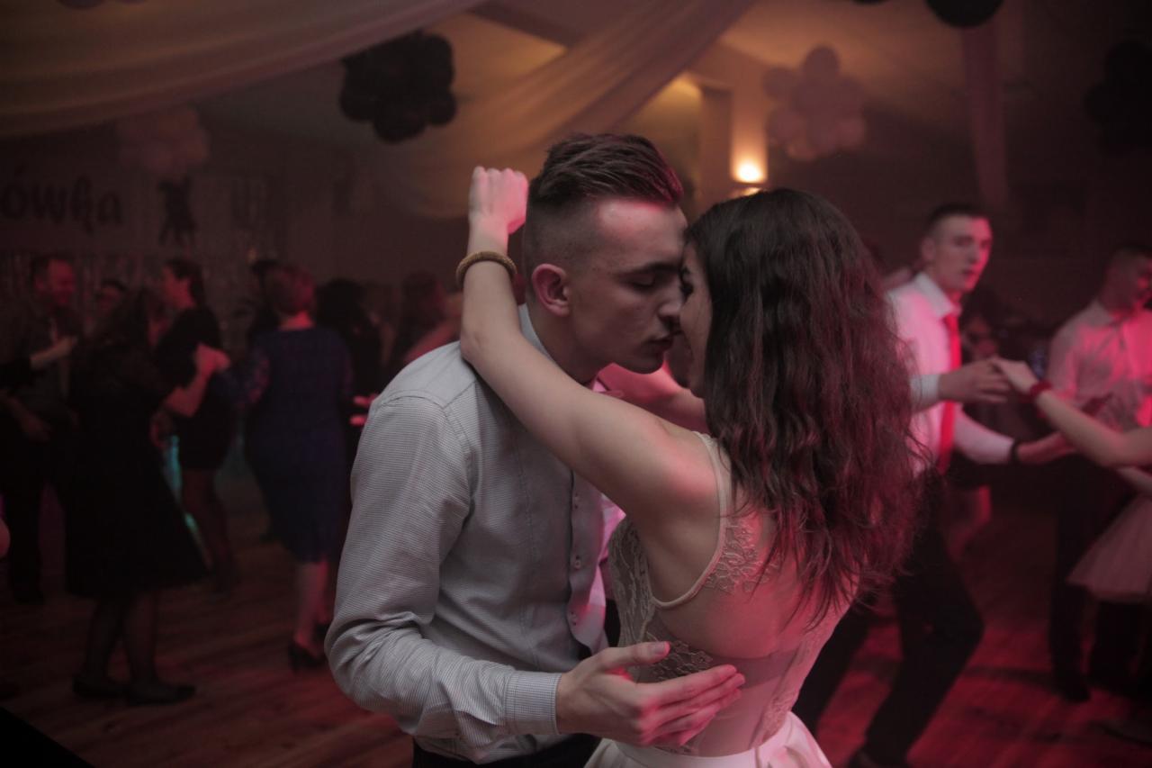 Para młoda w objęciach podczas pierwszego tańca na sali balowej z parkietem i innymi gośćmi w tle, oświetlenie sceniczne w odcieniach czerwieni.