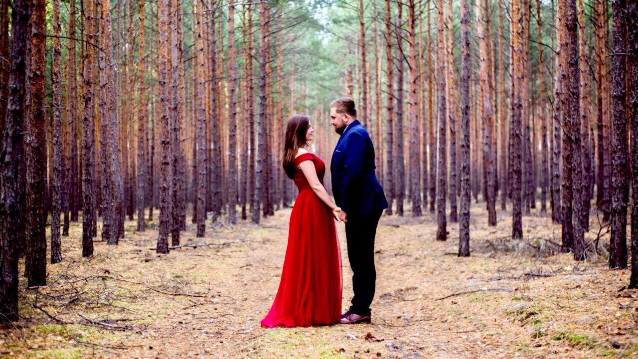 Para młoda w lesie sosnowym, ona w długiej czerwonej sukni, on w granatowym garniturze, trzymają się za ręce, patrząc na siebie.
