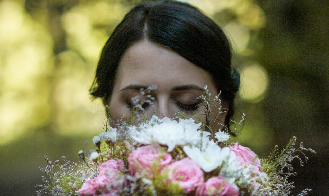 Panna młoda z zamkniętymi oczami, wąchająca bukiet z różowych róż i białych kwiatów na rozmytym zielonym tle.