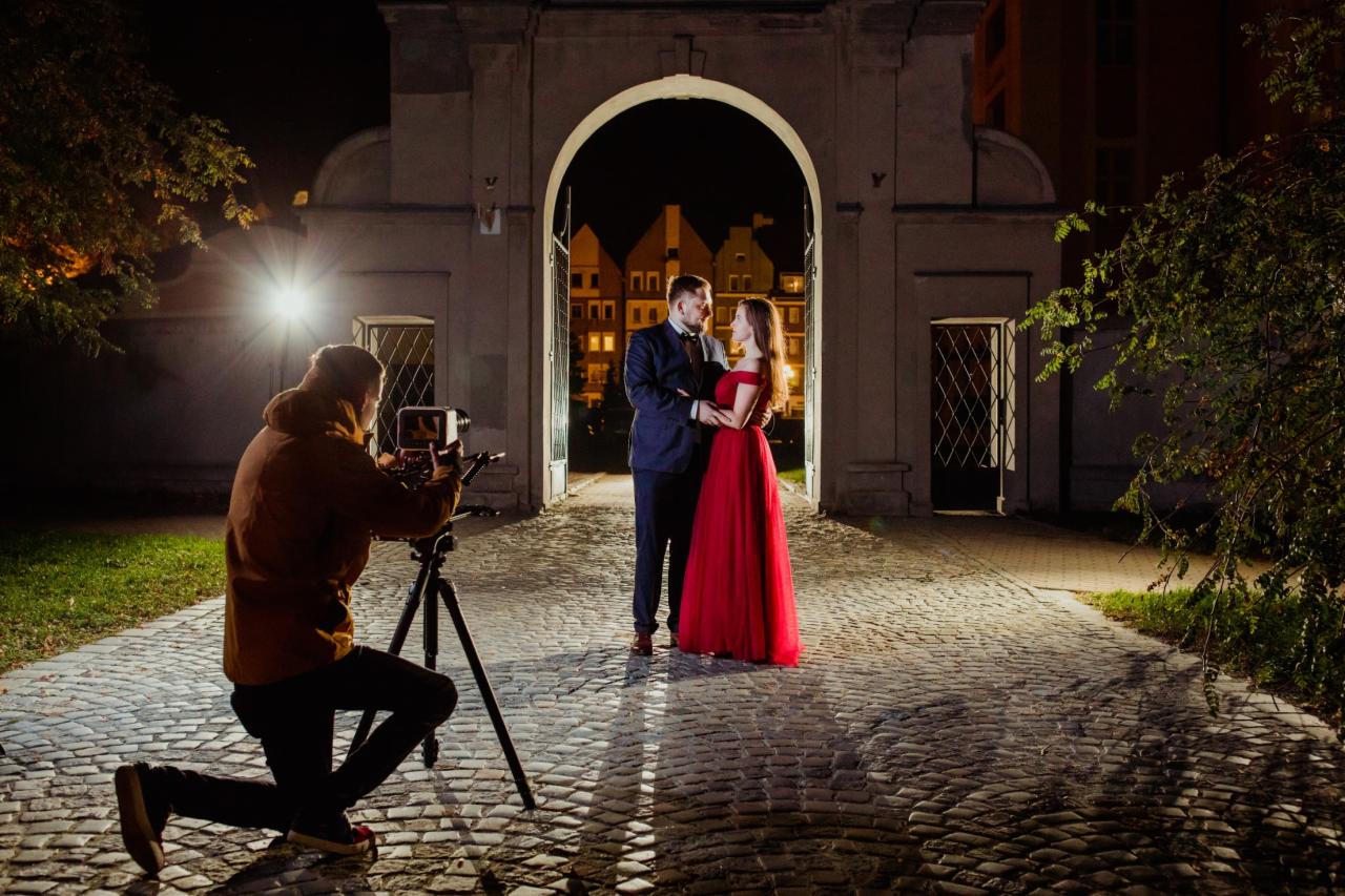 Kamerzysta na kolanach filmuje parę w strojach wieczorowych pod kamiennym łukiem, z widocznym oświetleniem i brukowaną nawierzchnią.
