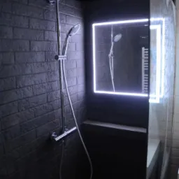 Nowoczesny prysznic z ciemną cegłą na ścianie, chromowaną armaturą i podświetlanym lustrem w kształcie prostokąta, odbijającym drugi prysznic.