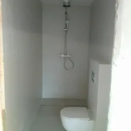 Wnętrze łazienki w trakcie prac wykończeniowych: szare płytki, wisząca toaleta, bateria prysznicowa z plastikowym pojemnikiem na górze, widoczne niedokończone fragmenty ścian.