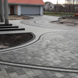 Ułożona kostka brukowa w odcieniach szarości z dekoracyjnymi obramowaniami, prowadząca do domu z kolumnami i schodami. Widoczny fragment trawnika i podjazd do garażu.