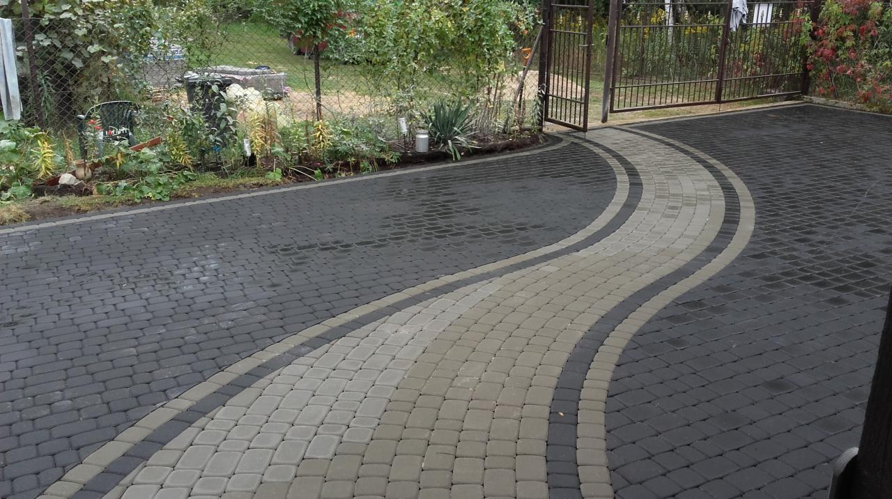 Ułożona kostka brukowa w kolorze grafitowym i szarym tworząca falujący wzór na podjeździe, widoczne ogrodzenie i roślinność w tle.