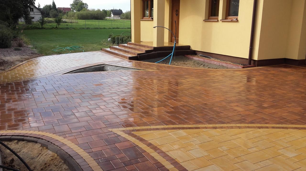 Świeżo ułożona, wilgotna kostka brukowa w odcieniach brązu i beżu tworzy wzór na podjeździe i ścieżce prowadzącej do domu z żółtą elewacją. Widoczne schody wejściowe i trawnik w tle.