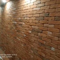 Ściana wykończona cegiełkami klinkierowymi w odcieniach czerwieni i brązu, oświetlona punktowo, z widocznymi fugami, ułożona na ścianie w rogu pomieszczenia.