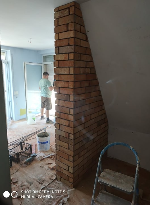 Wznoszenie dekoracyjnej kolumny z cegły klinkierowej wewnątrz pomieszczenia, widoczny pracownik w tle oraz drabina i narzędzia.