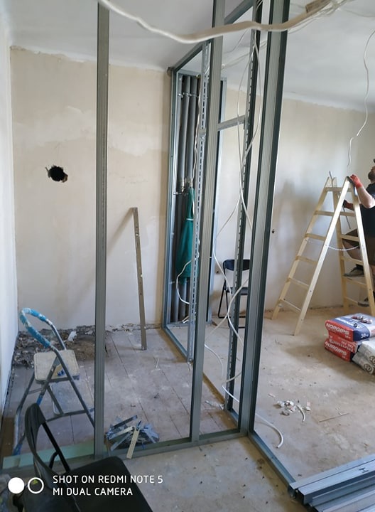 Pokój w trakcie remontu: widoczne metalowe profile ścianek działowych, wystające przewody elektryczne, drabina, worki z zaprawą oraz częściowo zerwana podłoga.