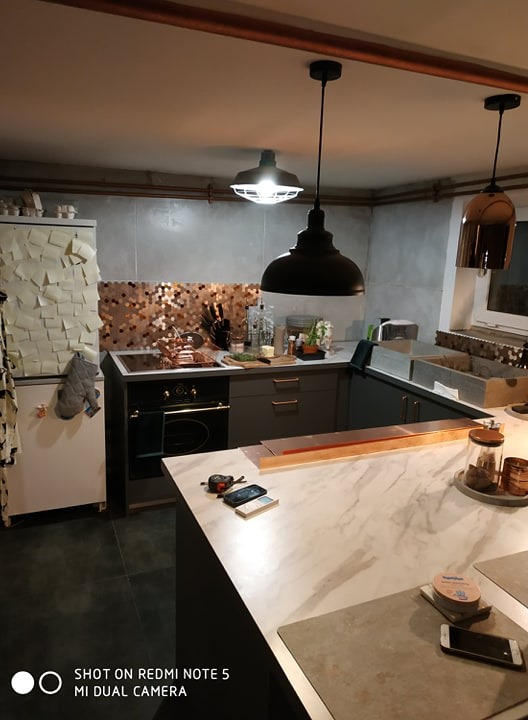 Kuchnia w trakcie remontu: widoczne szare szafki, miedziana mozaika nad blatem, trzy lampy wiszące w stylu industrialnym, rury miedziane pod sufitem, lodówka z naklejonymi kartkami, oraz narzędzia...