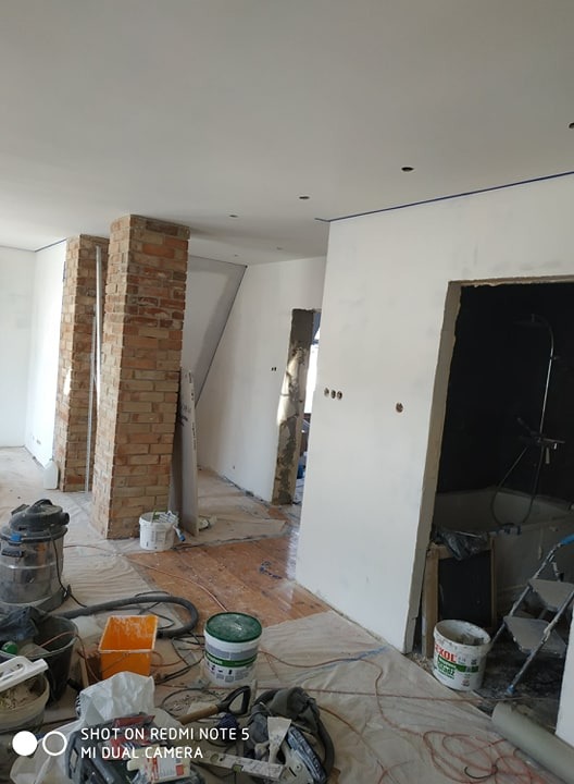 Wnętrze w trakcie prac remontowych: świeżo pomalowane białe ściany z niebieską taśmą malarską przy suficie, odsłonięte ceglane filary, zabezpieczona podłoga i widoczne narzędzia oraz pojemniki...