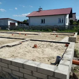 Fundamenty domu jednorodzinnego w trakcie budowy, widoczne bloczki betonowe, piasek, rury kanalizacyjne w wykopach, w tle dom z czerwoną dachówką i błękitne niebo.