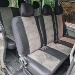 Wnętrze busa z trzema rzędami foteli w odcieniach szarości i czerni, widoczne pasy bezpieczeństwa, gumowa wycieraczka na podłodze, widok przez okno na zieleń.