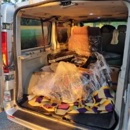 Wnętrze srebrnego busa załadowane do przeprowadzki: meble owinięte folią, rower zabezpieczony na kolorowej narzucie w tylnej części pojazdu, widok przez otwarte drzwi na zielone pole.