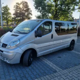 Srebrny van Renault Trafic z czarnymi felgami zaparkowany na brukowanym parkingu w słoneczny dzień, z widocznym odbiciem drzew w szybach.