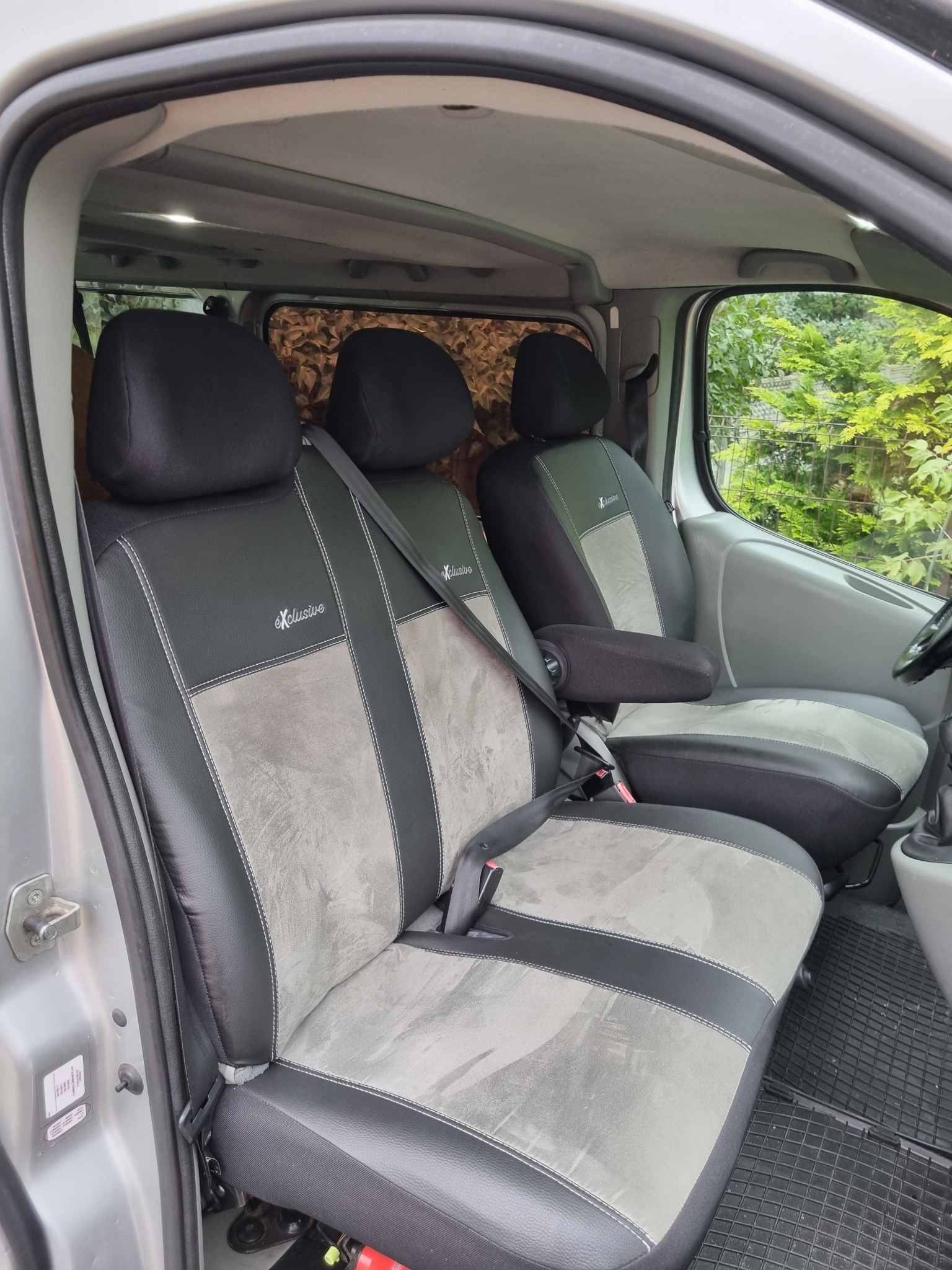 Wnętrze srebrnego vana z trzema fotelami w czarno-szarych pokrowcach z napisem 'Exclusive', widok z otwartych drzwi na fotele i zielone drzewa za oknem.