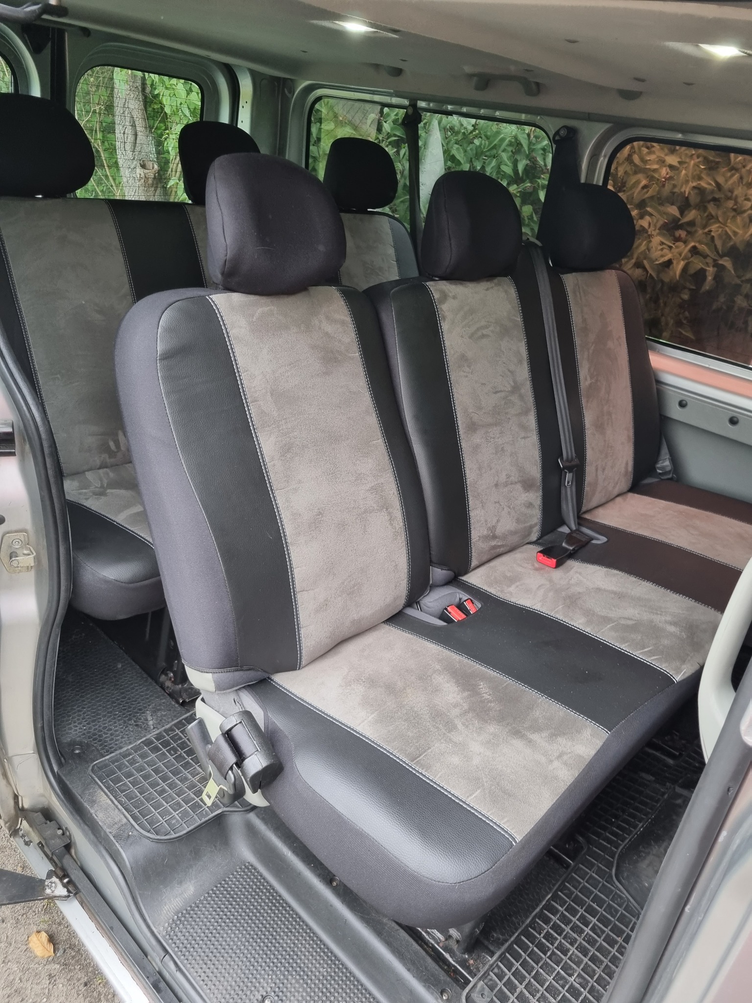 Wnętrze busa z trzema rzędami foteli w odcieniach szarości i czerni, widoczne pasy bezpieczeństwa, gumowa wycieraczka na podłodze, widok przez okno na zieleń.