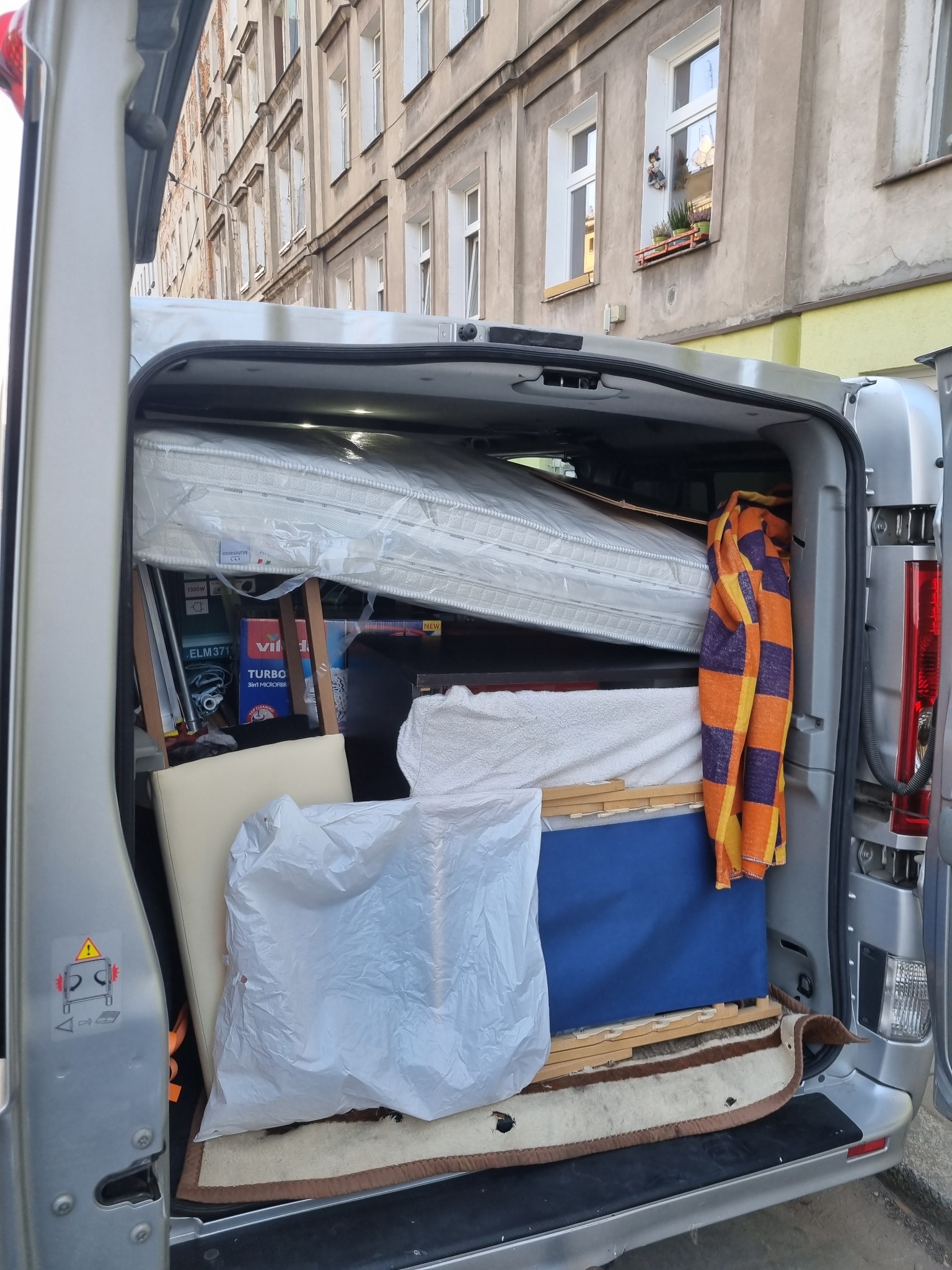 Wnętrze srebrnego vana załadowane do przeprowadzki: materac w folii, krzesło, kartony, koc w pomarańczowo-fioletową kratę, widok na budynek mieszkalny w tle.