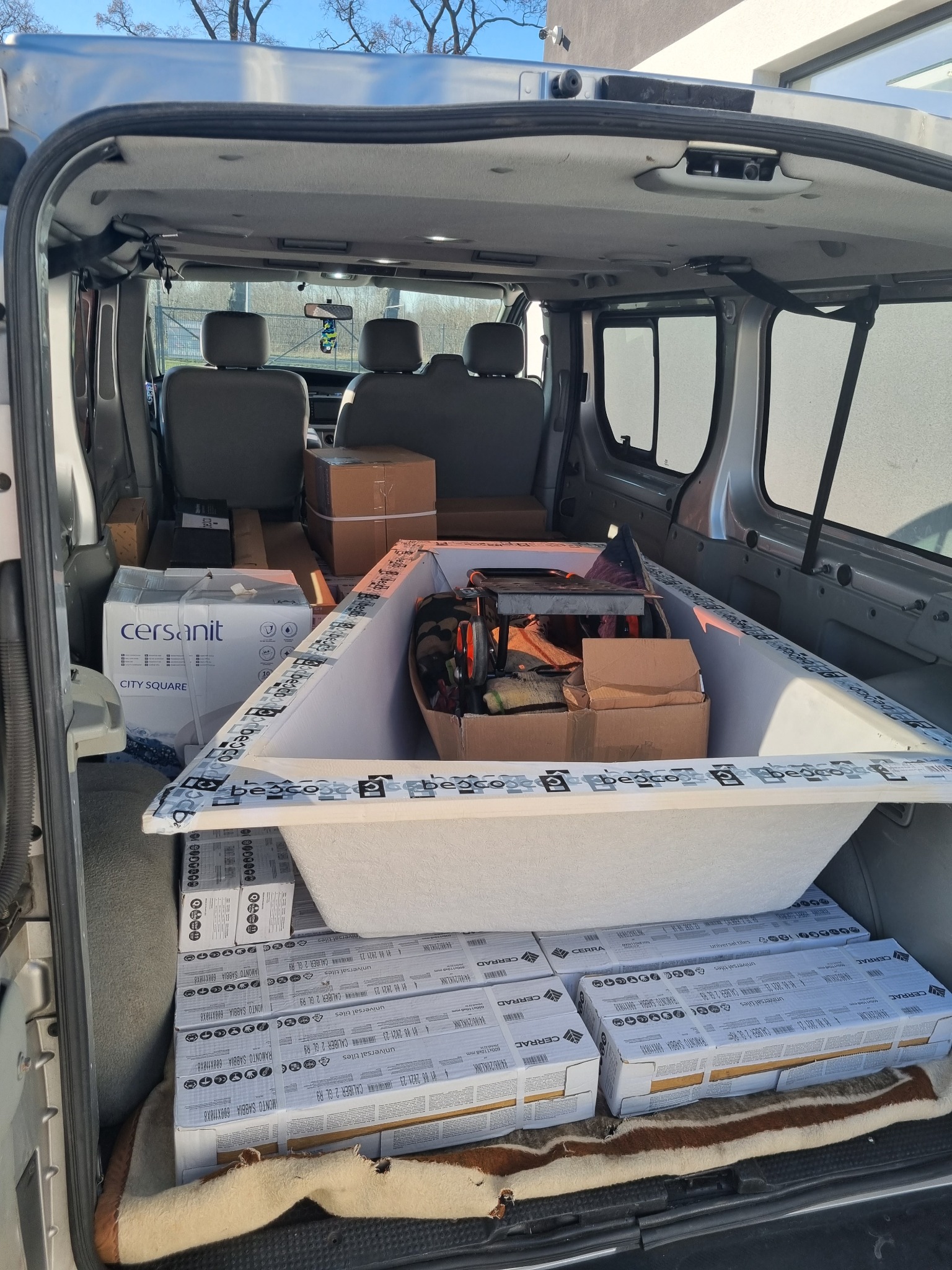 Wnętrze srebrnego vana załadowane materiałami budowlanymi, w tym nową białą wanną Besco, kartonami z płytkami Cersanit City Square i Cerad, oraz narzędziami w kartonach.