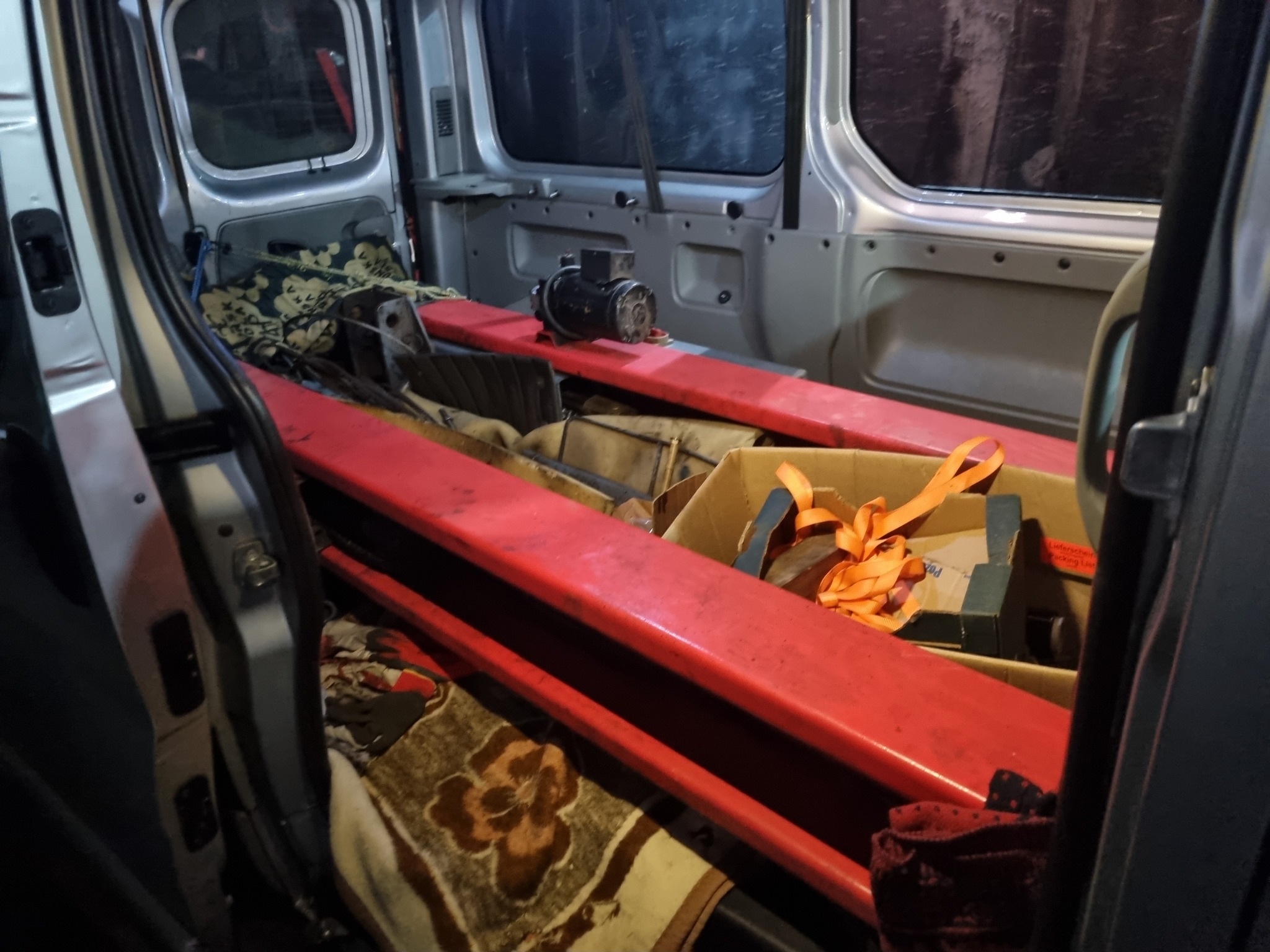 Wnętrze srebrnego busa z otwartymi drzwiami, załadowane czerwonymi, metalowymi belkami, kartonami i kocami w nieładzie, widoczny silnik elektryczny na belce, oświetlenie wnętrza słabe.