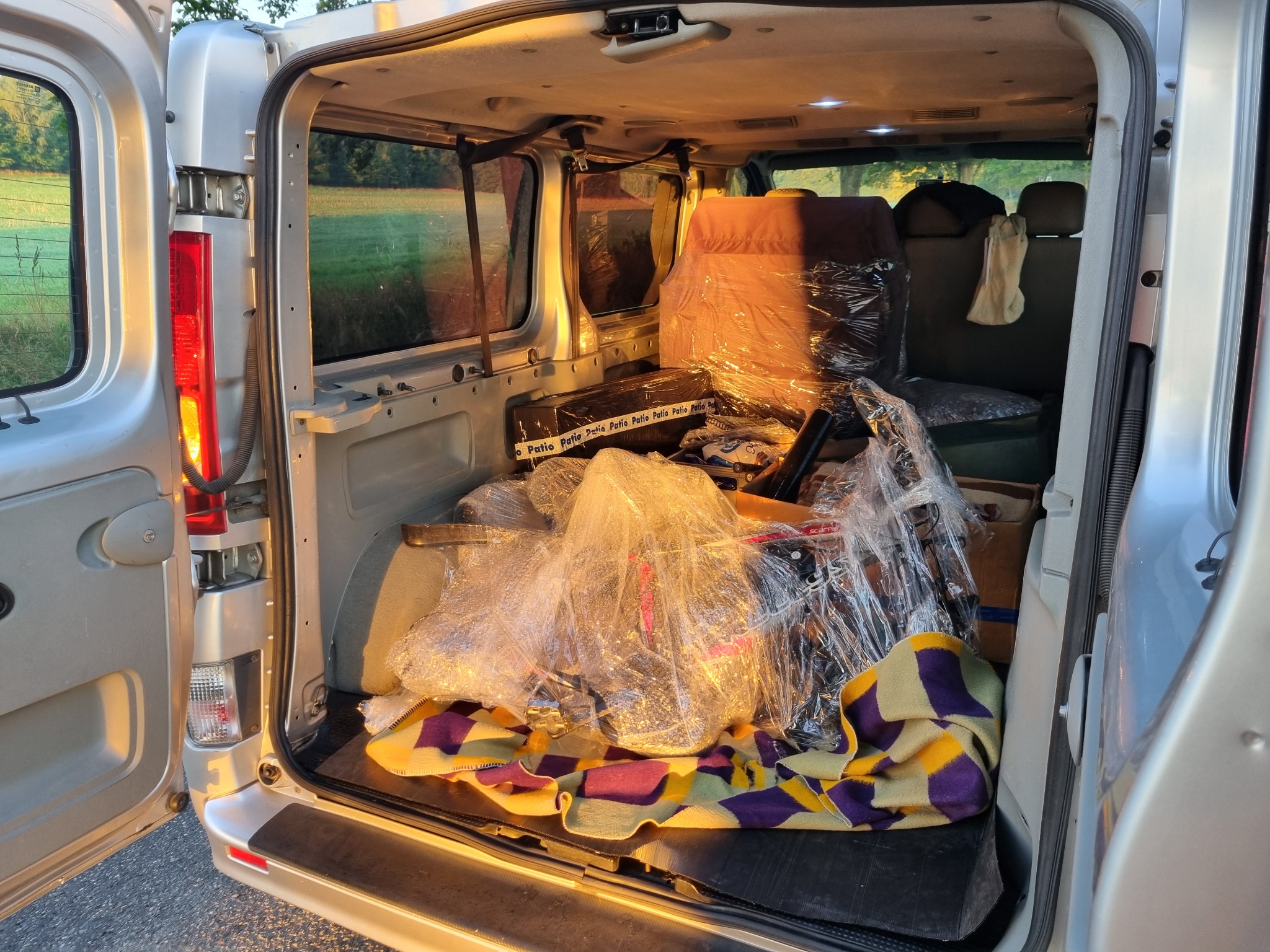 Wnętrze srebrnego busa załadowane do przeprowadzki: meble owinięte folią, rower zabezpieczony na kolorowej narzucie w tylnej części pojazdu, widok przez otwarte drzwi na zielone pole.