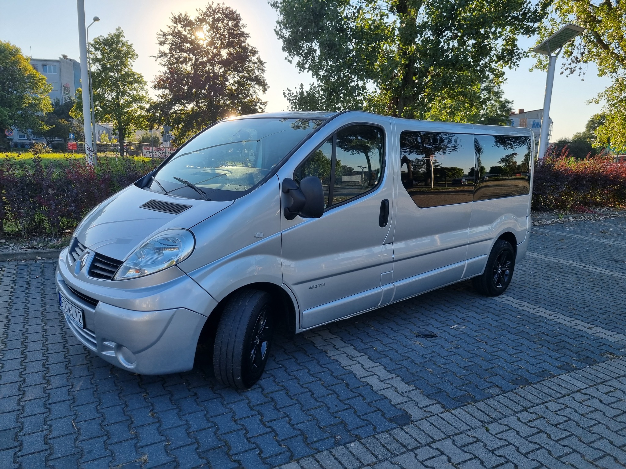 Srebrny van Renault Trafic z czarnymi felgami zaparkowany na brukowanym parkingu w słoneczny dzień, z widocznym odbiciem drzew w szybach.