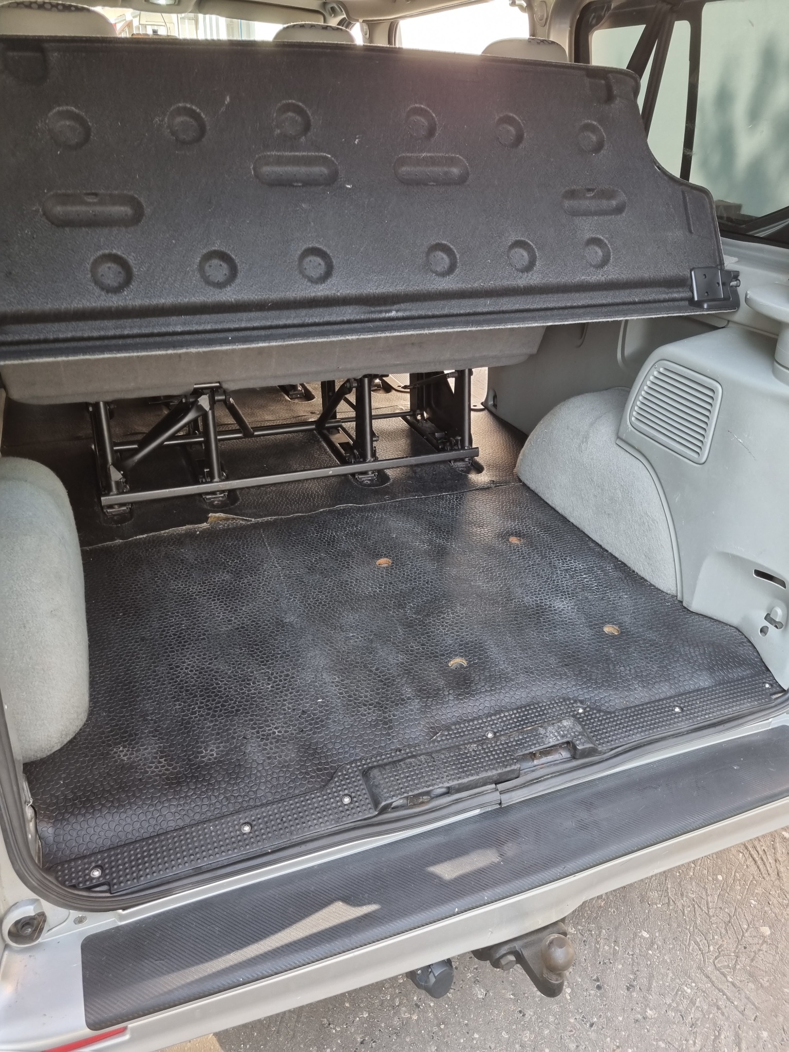 Wnętrze bagażnika samochodu typu van z podniesioną tylną półką, widoczna gumowa podłoga z okrągłymi otworami i hak holowniczy na zewnątrz.