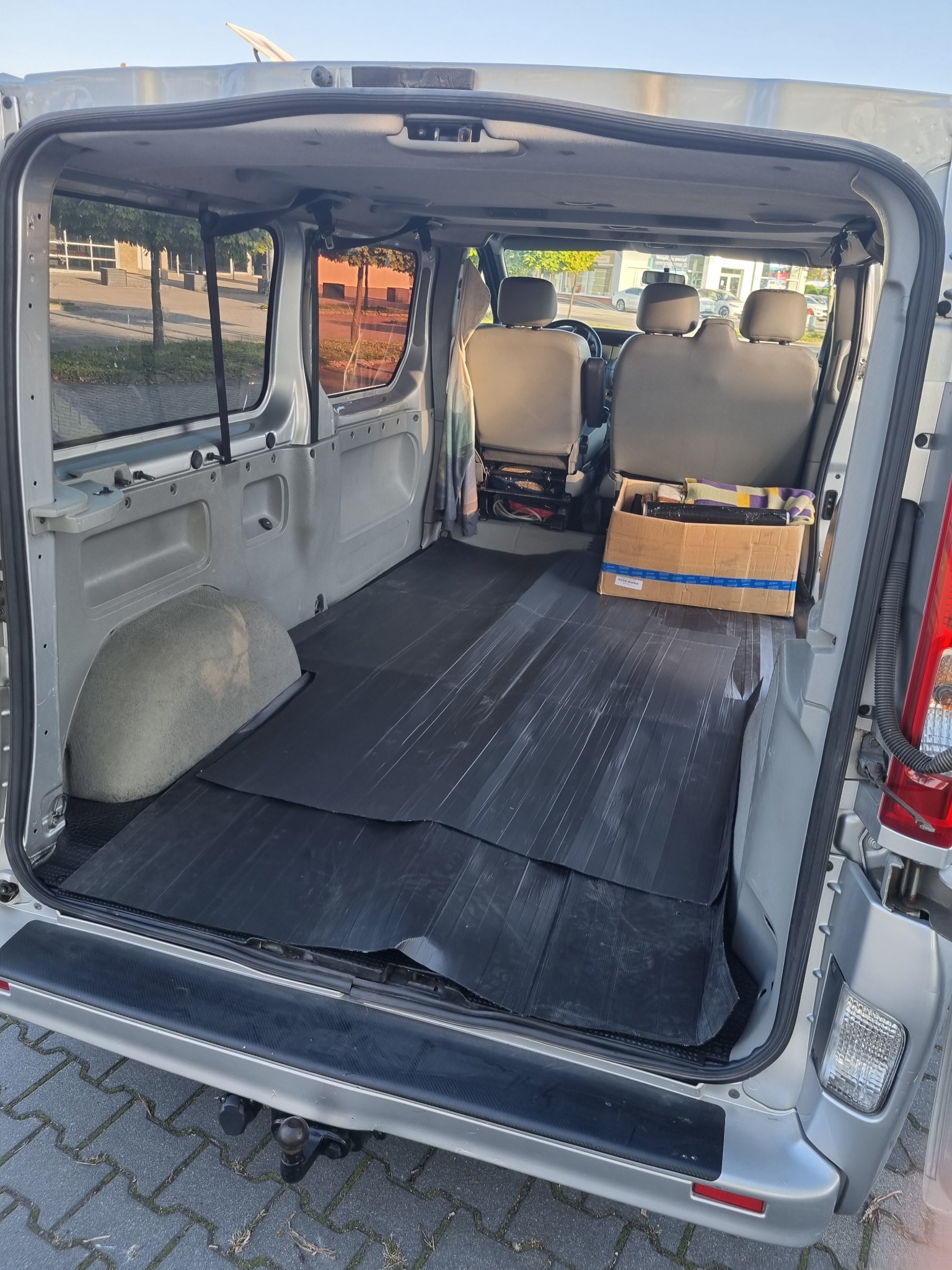 Wnętrze srebrnego busa z otwartymi tylnymi drzwiami, widoczna pusta przestrzeń ładunkowa wyłożona czarną gumową matą oraz karton z przedmiotami z tyłu, trzy rzędy siedzeń, hak holowniczy.