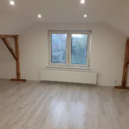 Poddasze po remoncie z drewnianymi belkami konstrukcyjnymi, jasną podłogą, białymi ścianami i oknem z widokiem na zewnątrz.