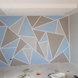 Ściana w pokoju pomalowana w geometryczny wzór z użyciem niebieskiego, szarego i białego koloru, widoczne puszki z farbą i taśma malarska na podłodze.